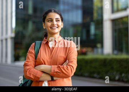 Jeune femme indienne portant un sac à dos et croisant les bras, regardant loin et souriant devant un bâtiment universitaire moderne, représentant l'éducation, la réussite et les opportunités futures Banque D'Images