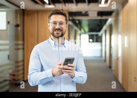 Homme confiant souriant directement à la caméra tout en tenant un smartphone, debout dans un couloir de bureau contemporain et lumineux en bois et verre Banque D'Images