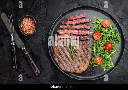 Steak de ribeye tranché juteux avec une riche surface marbrée et brunie. Côtelette de bœuf grillée servie avec salade sur une assiette en céramique noire Banque D'Images