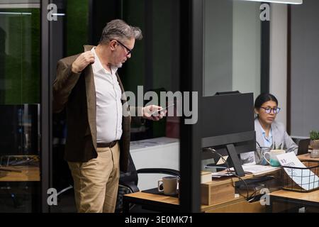 Homme caucasien d'âge moyen entrant au bureau tout en tenant une veste et en regardant une tablette, jeune femme hispanique assise au bureau travaillant sur ordinateur dans un espace de travail moderne Banque D'Images