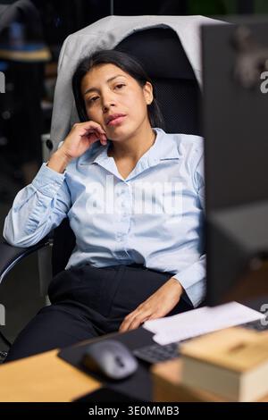 Portrait d'une femme hispanique assise au bureau se penchant en arrière dans une chaise de bureau reposant la tête sur la main regardant dans la caméra semblant réfléchie pendant la journée de travail Banque D'Images