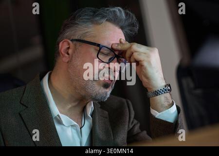 Homme caucasien d'âge moyen portant des lunettes assis au bureau touchant le front avec la main, paraissant stressé ou profond dans la pensée, travaillant tard dans l'environnement de bureau Banque D'Images