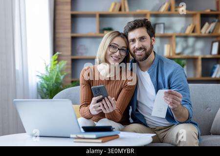 Couple examinant les documents et les informations numériques sur un ordinateur portable et un smartphone, calculant les dépenses et les revenus communs, planifiant leurs économies futures ensemble Banque D'Images