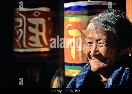 Une femme âgée se tient devant des roues à prières bouddhistes colorées au Bhoutan. Elle sourit tout en réfléchissant sur ses traditions et sa culture. Banque D'Images