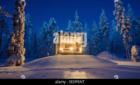 Nichée dans le paysage enneigé serein d'Akaslompolo, une cabane moderne brille chaleureusement sur fond de grands arbres givrés sous un ciel étoilé. Une retraite hivernale parfaite. Banque D'Images