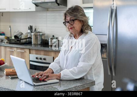 Femme âgée utilisant un ordinateur portable dans sa cuisine domestique, multitâche et restant connectée Banque D'Images
