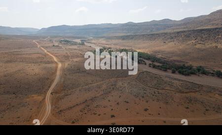 Vue aérienne d'une vallée désertique aride avec un chemin de terre sinueux, lit de rivière sec bordé d'arbres verts, et montagnes rocheuses lointaines sous ciel brumeux. Sokotr Banque D'Images
