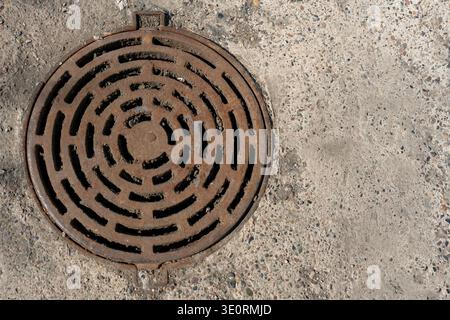Couvercle de trou d'homme rond rouillé avec motif de grille concentrique placé dans la chaussée en béton altéré, mettant en évidence la texture, l'infrastructure urbaine, la pourriture et l'ut Banque D'Images