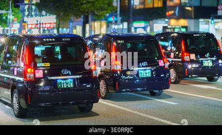 Tokyo, Japon - 14 octobre 2024, Une vue arrière panoramique de trois taxis noirs avec leurs feux arrière allumés, conduisant le long d'une rue la nuit dans le Ginza dist Banque D'Images