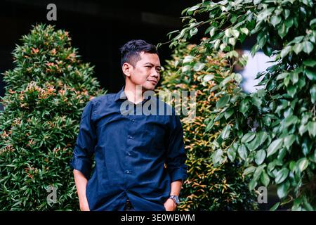 Un jeune homme asiatique dans une chemise à col bleu foncé se tient réfléchi, regardant loin au milieu d'un feuillage vert luxuriant et rougeâtre dans un cadre de jardin extérieur. Banque D'Images