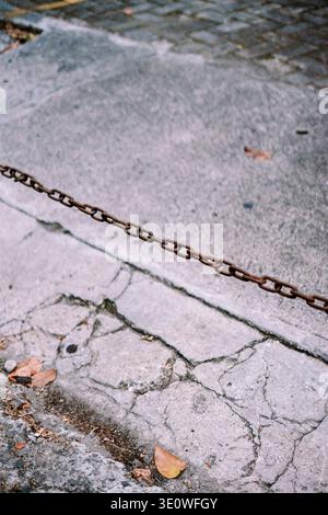 Gros plan d'une chaîne métallique rouillée et endommagée tendue sur un sol en béton fissuré et texturé, avec des feuilles sèches éparpillées. Banque D'Images