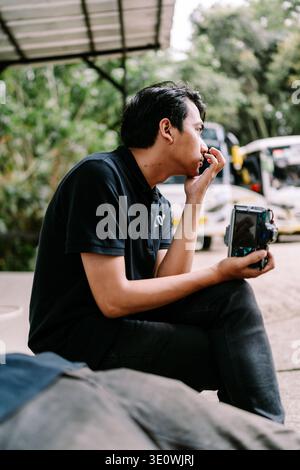 Un jeune homme asiatique réfléchi est assis à l'extérieur dans un cadre décontracté, se reposant avec un appareil photo à la main, regardant contemplatif. Banque D'Images