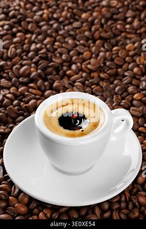Petite tasse à espresso blanche avec éclaboussures et gouttes, débordant de café aromatique reposant sur le lit de grains torréfiés Banque D'Images