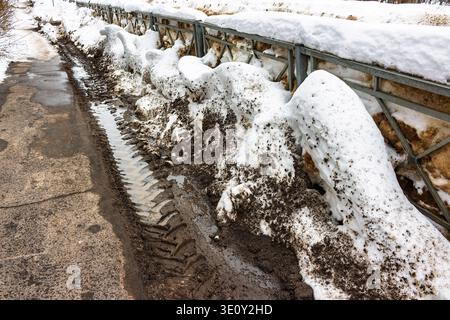 Des dérives sales de neige fondante bordent une clôture, des chenilles profondes du tracteur coupent la neige fondante boueuse. Une scène vivante de dégel de fin d'hiver Banque D'Images
