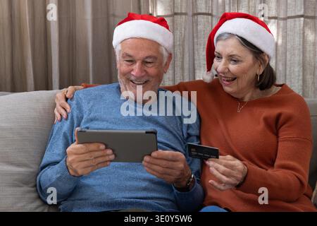 Couple senior assis sur le canapé dans le salon portant des chapeaux de Père Noël en utilisant la tablette, carte de crédit. Personnes âgées, détente, technologie, festif, confortable, intimité, convivialité || autorisation du modèle Banque D'Images