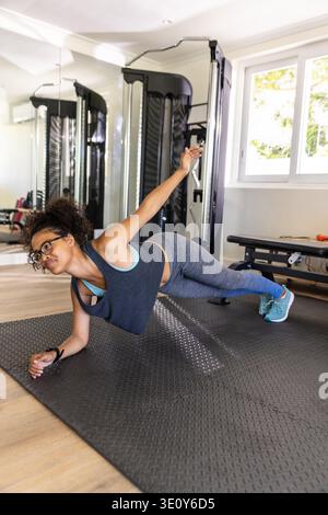 Femme afro-américaine exécutant planche latérale sur tapis texturé noir dans la salle de gym à domicile avec machine à câble. Fitness, force, bien-être, entraînement, mode de vie actif, aérobie, moderne || autorisation du modèle Banque D'Images