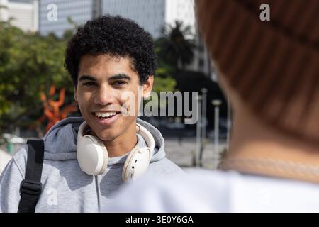 Adolescent afro-américain et ami souriant et parlant dans Urban plaza, portant un sweat à capuche et des écouteurs. Jeunesse, étudiant, conversation, candide, streetwear, sac à dos, bonnet || autorisation du modèle Banque D'Images