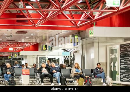 Passagers attendant à la porte d'embarquement dans le terminal moderne de l'aéroport avec coin salon, panneaux numériques et structure de plafond en acier rouge Banque D'Images