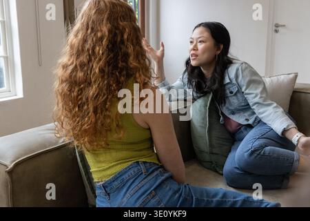 Deux femmes assises sur un canapé beige dans le salon parlant et faisant des gestes avec un oreiller vert Banque D'Images