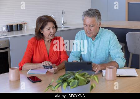 Couple senior vérifiant la tablette à la table de la cuisine, femme en blouse rouge tenant la carte de crédit Banque D'Images
