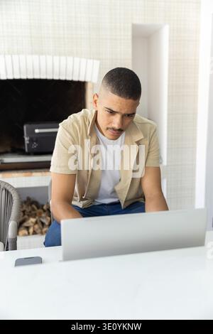 Homme dans ses 30 ans assis à la maison à l'aide d'un ordinateur portable, smartphone gauche, portant une chemise beige, cheminée derrière Banque D'Images