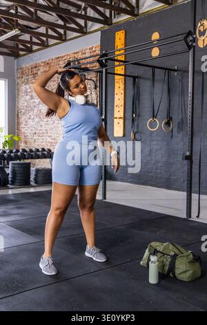 Femme afro-américaine adulte étirant le cou dans un studio de fitness portant une tenue bleue près de la plate-forme de pull-up Banque D'Images