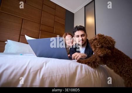 Jeune couple souriant à l'ordinateur portable avec caniche dans la chambre Banque D'Images