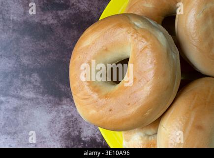 Vue de dessus d'un groupe de bagels simples sur une grande plaque jaune au sommet d'une table marbrée rouge illuminée d'un éclairage naturel. Banque D'Images