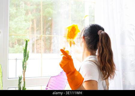Jeune brune lave la fenêtre dans des gants et un tablier, nettoyage de la maison, plante en pot sur la fenêtre, cuisine, intérieur lumineux, promotion de l'entreprise de nettoyage, verre c Banque D'Images