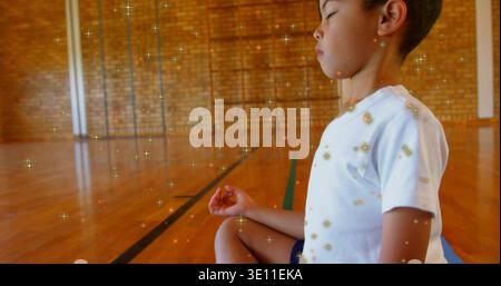 Enfant assis en T-shirt blanc short sombre méditant sur tapis dans la salle de gym, superposition scintillante, espace de copie Banque D'Images