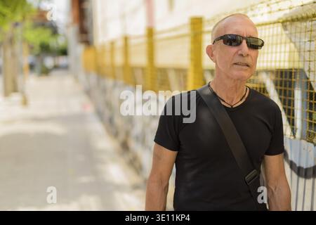 Senior homme chauve touristique pensée tout en portant des lunettes de soleil contre le mur de graffitis urbains sale dans la rue publique à l'extérieur Banque D'Images