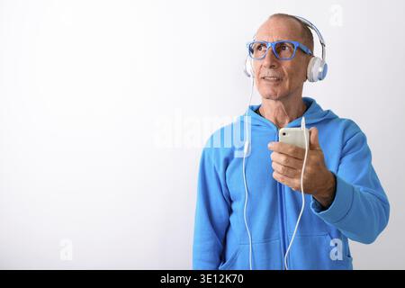 Studio shot of bald senior man holding mobile phone alors que penser et d'écouter de la musique avec des lunettes contre fond blanc Banque D'Images