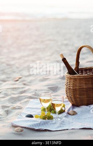 Deux verres à vin avec du vin blanc debout sur le sable, sur la plage, à côté de raisins et panier en osier avec bouteille de vin. Les vagues de la mer en arrière-plan Banque D'Images