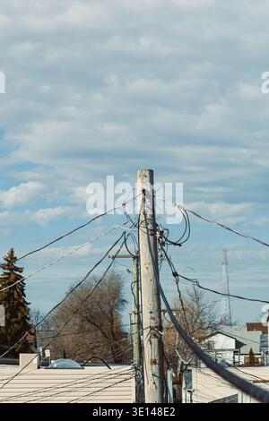 Un poteau utilitaire en bois se trouve dans un quartier avec de nombreux fils enchevêtrés autour d'elle. Le fond montre des maisons et une tour contre le ciel nuageux. Banque D'Images