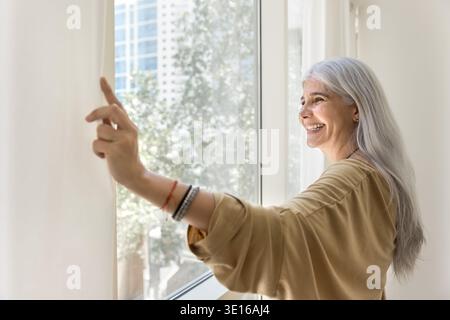 Souriante femme âgée ouvrant le rideau tout en se tenant près de la fenêtre Banque D'Images