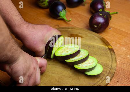 Vue rapprochée des mains masculines coupant des aubergines sur une planche à découper dans la cuisine Banque D'Images
