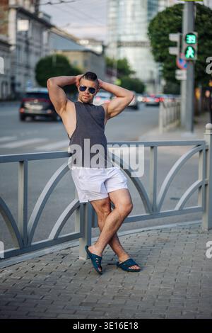 Jeune homme musclé avec des lunettes de soleil posant près d'un feu de circulation dans une rue de la ville, portant un débardeur et un short Banque D'Images