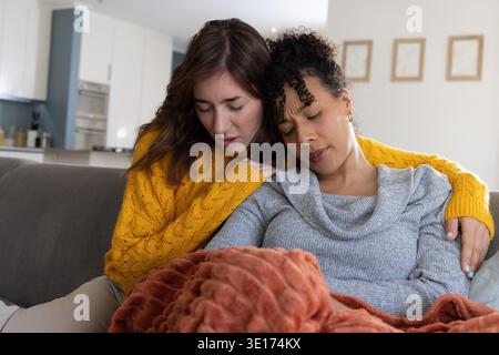Diverses femmes assises sur un canapé gris en moutarde et chandails gris clair, partageant une couverture orange. Couple, confortable, intime, maison, canapé, cuisine, texturée || autorisation du modèle Banque D'Images