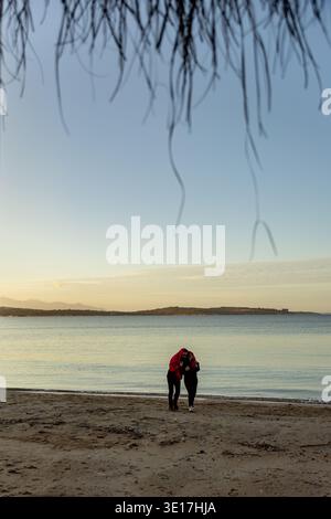 Un couple profite d'un moment serein en marchant le long d'une plage de sable, enveloppés dans les bras de l'autre sous un ciel dégagé. La mer s'étend tranquillement à l'arrière Banque D'Images