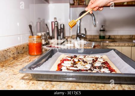Un cuisinier fait couler du miel sur une tarte avec une trempette de miel dans une cuisine moderne. La tarte, garnie d'un mélange de fruits secs et de noix, repose sur un parchmen Banque D'Images