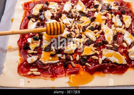 Un gros plan d'une tarte salée en cours de préparation, avec des couches de fromage, de fruits secs, de pignons de pin et de miel sur une base de pâte feuilletée, prête pour la cuisson. Banque D'Images