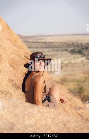 Une femme est assise sur une surface rocheuse ensoleillée portant un chapeau brun, profitant de la vaste vue panoramique sur le paysage désertique de Namibie. La Rock Arch forme un stunnin Banque D'Images