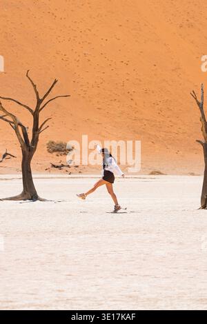 Une femme marche joyeusement entre deux anciens arbres sans feuilles à Deadvlei, en Namibie. Le contraste saisissant du paysage désertique aride met en évidence son adv Banque D'Images