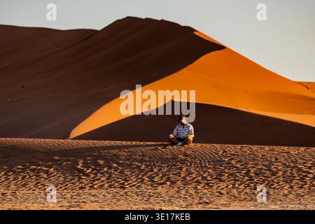 Un homme est assis calmement sur le sable ondulé des dunes de Sossusvlei, en Namibie, avec une lumière dorée soulignant le vaste et majestueux paysage désertique ondulé Banque D'Images