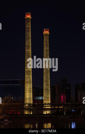 Deux hautes cheminées industrielles, éclairées par une lumière chaude, se dressent bien en évidence contre le ciel nocturne, contrastant avec l'horizon i moderne de Pékin Banque D'Images