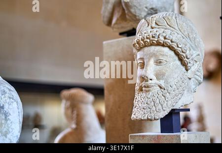 Vue rapprochée d'une ancienne sculpture en pierre représentant une tête barbu d'homme avec des cheveux bouclés détaillés et un bandeau décoratif ou une couronne, affichée sur un Banque D'Images