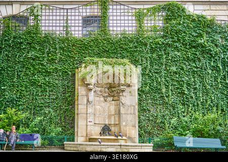 Fontaine en pierre historique avec bâtiment couvert de lierre dans le parc parisien, homme âgé sur banc, pigeons, scène urbaine tranquille Banque D'Images