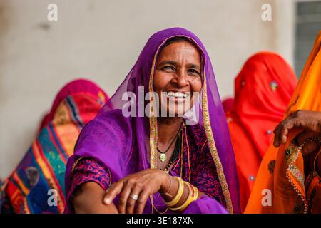 Une femme souriante dans un sari violet et des bracelets jaunes s'assoit avec d'autres personnes habillées de couleurs vives à Pushkar Mela. Banque D'Images