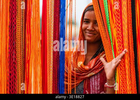 Une jeune femme souriante à Pushkar Mela pose derrière un rideau dense de cordes tressées rouges, jaunes et bleues éclatantes. Banque D'Images