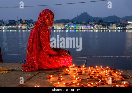 Une jeune femme dans un sari rouge est assise près du lac Pushkar, Pushkar Sarovar, entourée de lampes à huile allumées avec la ville illuminée visible au loin. Banque D'Images